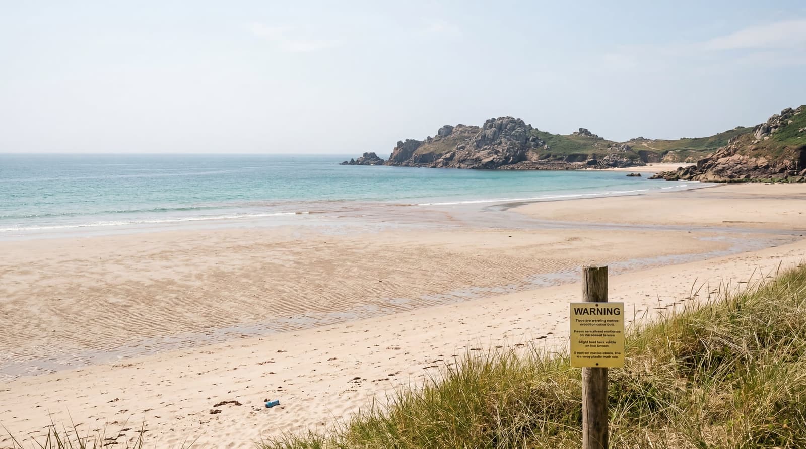 A wide, empty sandy bay on Jersey's north coast at low tide under bright, hazy summer sunlight, a small yellow warning notice on a wooden post in the foreground near the dune line
